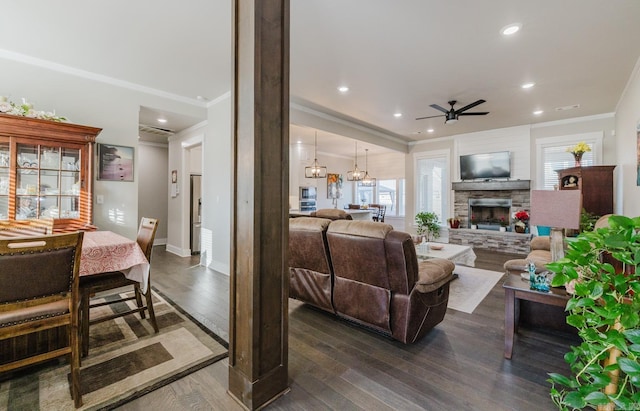 living area featuring dark wood-style floors, ornamental molding, ceiling fan with notable chandelier, a stone fireplace, and recessed lighting