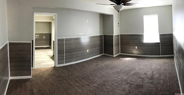 empty room featuring dark colored carpet, wainscoting, a ceiling fan, and wooden walls