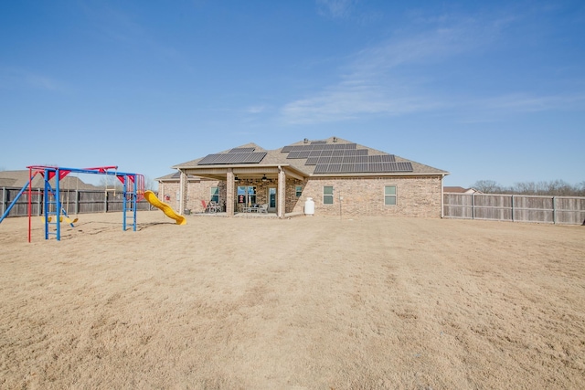 back of house with a fenced backyard, roof mounted solar panels, a playground, and brick siding