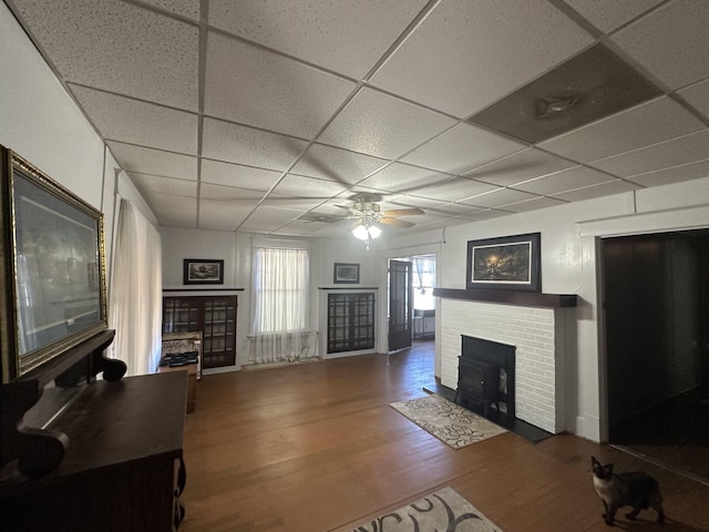 living room featuring a drop ceiling, hardwood / wood-style floors, a brick fireplace, and ceiling fan