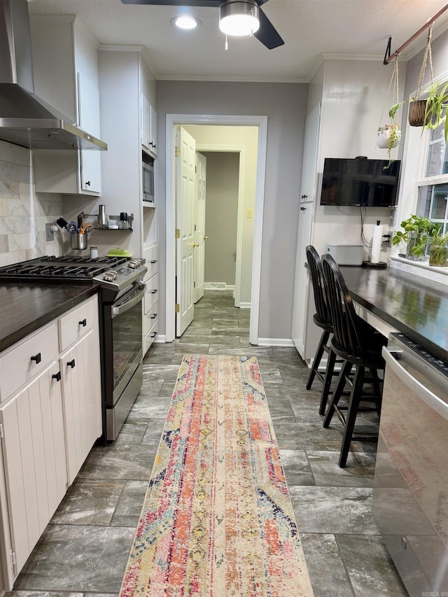 kitchen with wall chimney exhaust hood, appliances with stainless steel finishes, ornamental molding, white cabinets, and backsplash