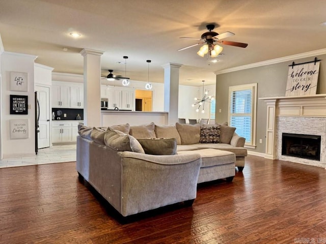 living room featuring a stone fireplace, decorative columns, ornamental molding, dark hardwood / wood-style flooring, and ceiling fan with notable chandelier