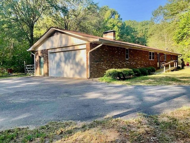 view of side of home featuring driveway, an attached garage, a chimney, a view of trees, and brick siding