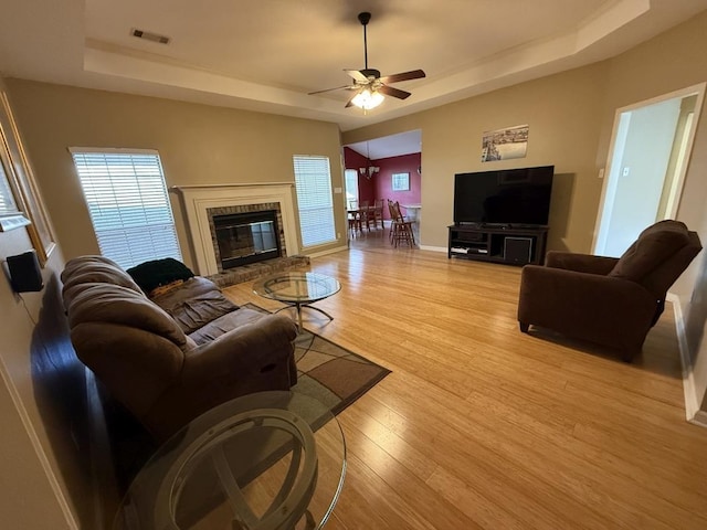 living room with a brick fireplace, a tray ceiling, ceiling fan, and light wood-type flooring