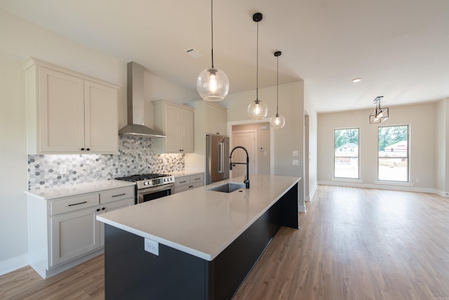 kitchen with wall chimney range hood, sink, stainless steel appliances, an island with sink, and decorative light fixtures