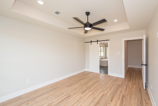 unfurnished bedroom with ensuite bath, ceiling fan, a tray ceiling, light hardwood / wood-style floors, and a barn door