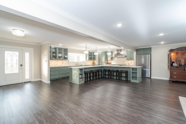 kitchen featuring pendant lighting, a kitchen island with sink, wall chimney range hood, and stainless steel fridge