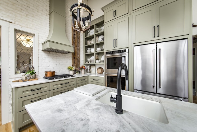 kitchen with sink, appliances with stainless steel finishes, light stone countertops, custom range hood, and green cabinetry