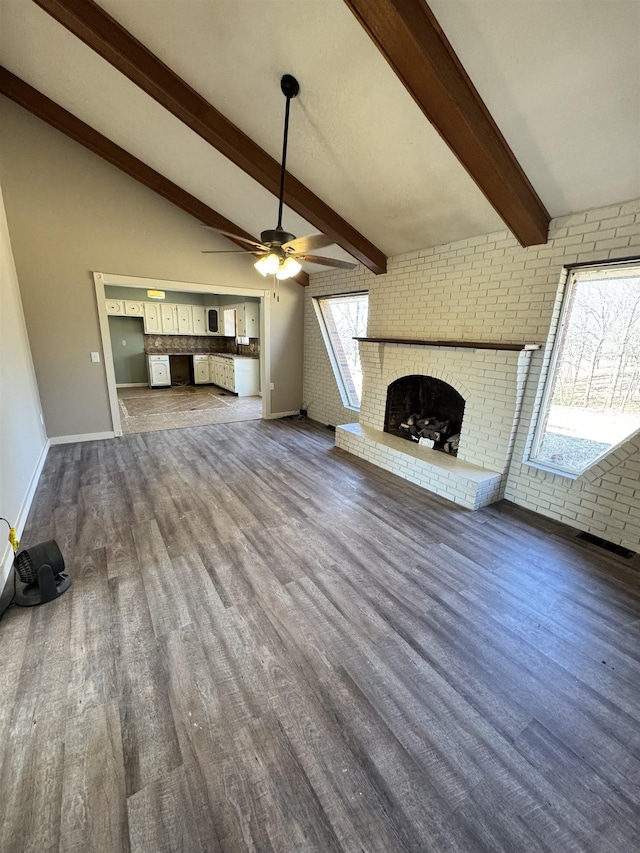 unfurnished living room featuring dark wood-type flooring, ceiling fan, a fireplace, lofted ceiling with beams, and brick wall