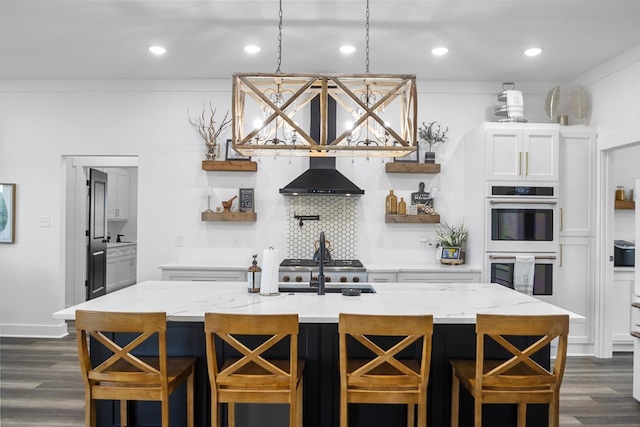 kitchen with wall chimney exhaust hood, double oven, a center island with sink, and white cabinets