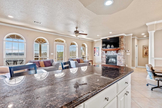 kitchen with crown molding, dark stone countertops, white cabinets, light tile patterned flooring, and a stone fireplace