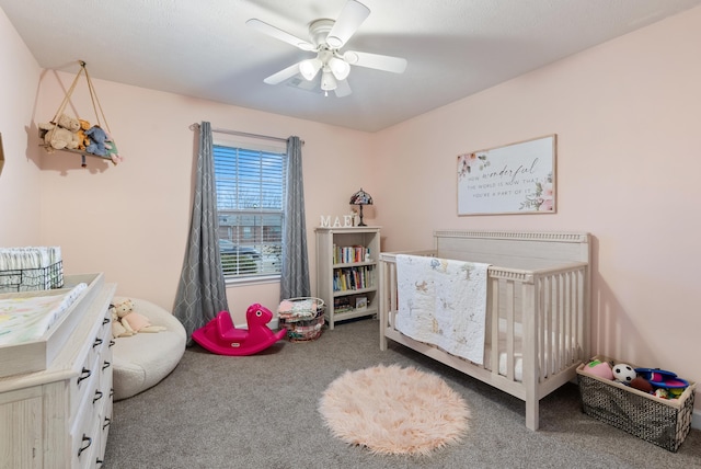 bedroom with a nursery area, ceiling fan, and dark colored carpet