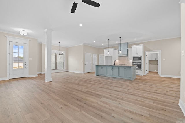 unfurnished living room with sink, decorative columns, ornamental molding, light hardwood / wood-style floors, and ceiling fan with notable chandelier