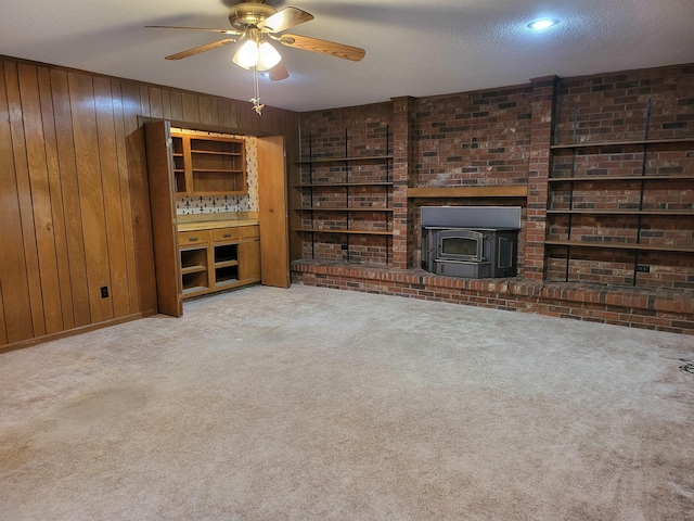 unfurnished living room featuring brick wall, wood walls, carpet floors, a wood stove, and ceiling fan