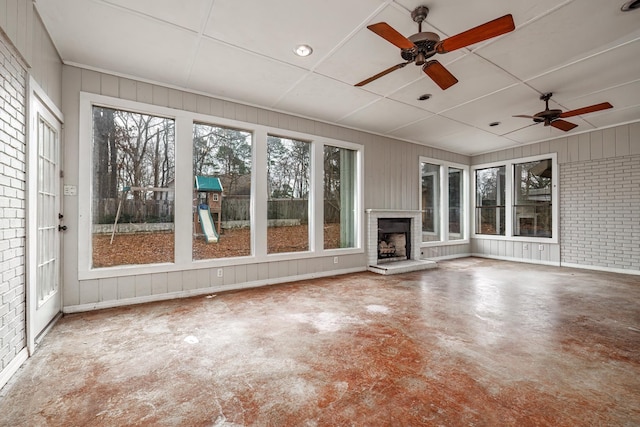 unfurnished living room with ceiling fan, brick wall, concrete flooring, and an outdoor fireplace