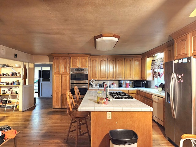 kitchen with wood-type flooring, a kitchen bar, a kitchen island with sink, tile counters, and stainless steel appliances