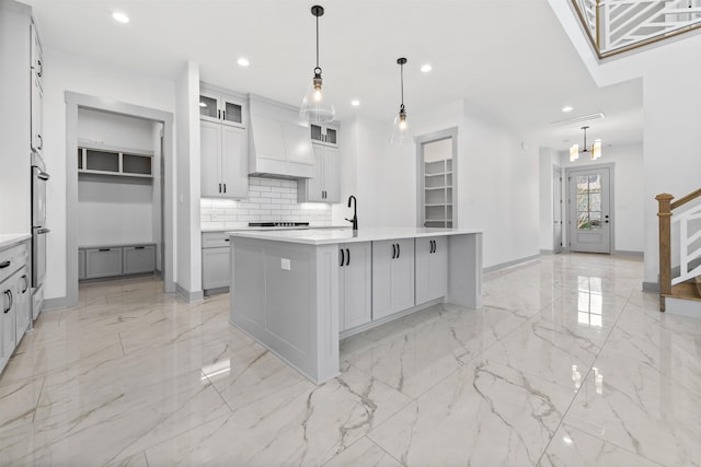 kitchen featuring double oven, decorative backsplash, hanging light fixtures, a kitchen island with sink, and custom range hood