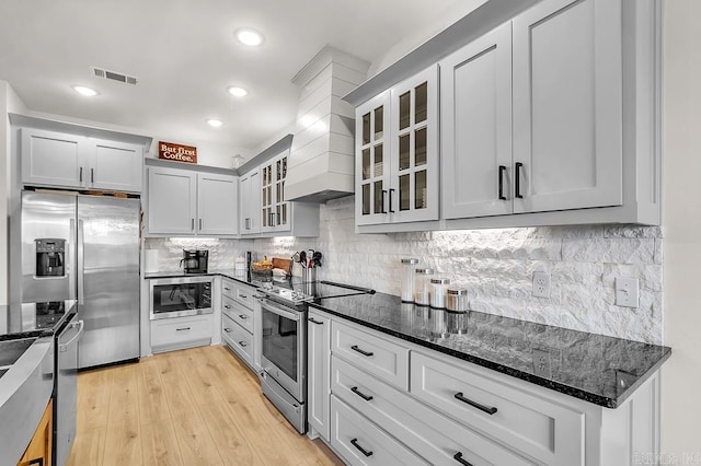 kitchen featuring custom exhaust hood, light wood-type flooring, dark stone counters, stainless steel appliances, and decorative backsplash