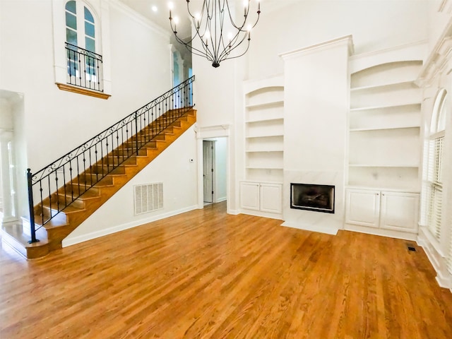 unfurnished living room with built in features, an inviting chandelier, a high ceiling, wood-type flooring, and ornamental molding