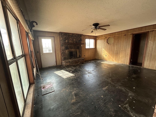 unfurnished living room with ceiling fan, wooden walls, a stone fireplace, and a textured ceiling