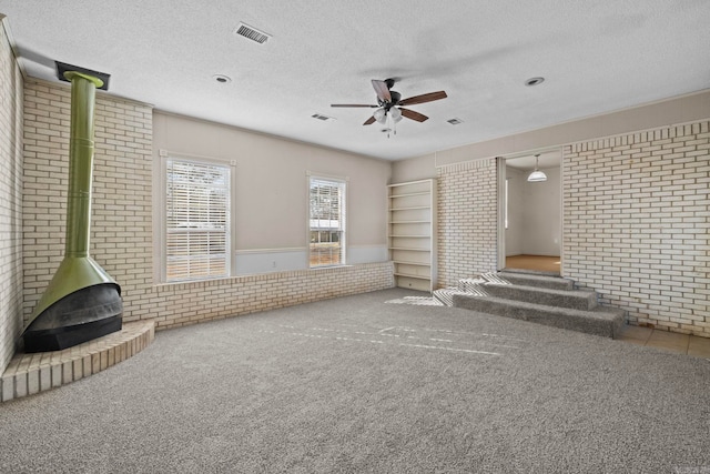 unfurnished living room with brick wall, a wood stove, carpet flooring, ceiling fan, and a textured ceiling