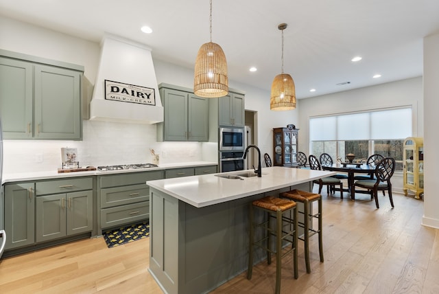 kitchen featuring premium range hood, appliances with stainless steel finishes, pendant lighting, sink, and a kitchen island with sink