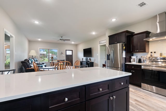 kitchen with appliances with stainless steel finishes, tasteful backsplash, ceiling fan, wall chimney range hood, and light hardwood / wood-style flooring