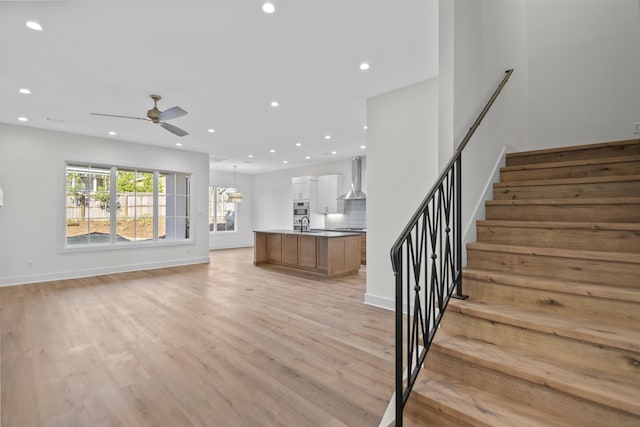 unfurnished living room with light wood-type flooring, stairway, ceiling fan with notable chandelier, and recessed lighting