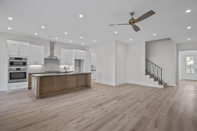 kitchen featuring open floor plan, a kitchen island with sink, a sink, wall chimney range hood, and white cabinetry