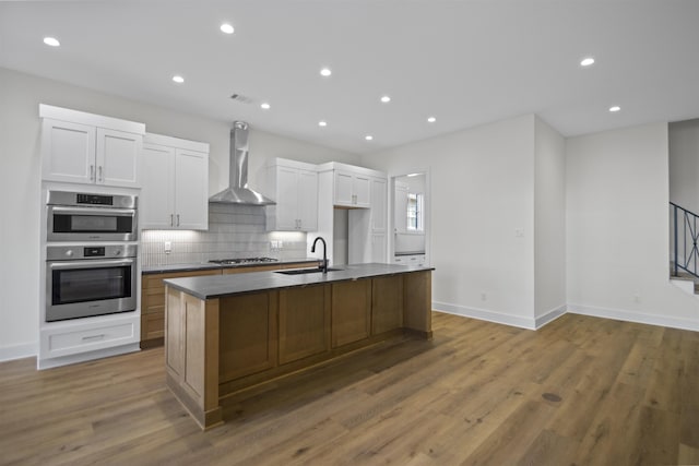 kitchen featuring appliances with stainless steel finishes, white cabinets, a sink, an island with sink, and wall chimney exhaust hood