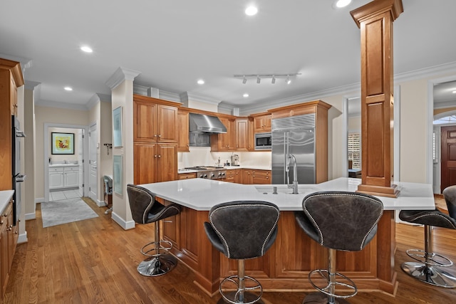 kitchen featuring a breakfast bar, decorative columns, built in appliances, wall chimney exhaust hood, and light hardwood / wood-style flooring