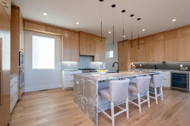 kitchen with wine cooler, light brown cabinetry, light stone counters, appliances with stainless steel finishes, and an island with sink