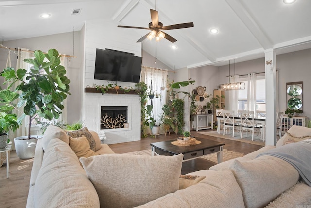 living room featuring lofted ceiling with beams, wood-type flooring, a large fireplace, and ceiling fan with notable chandelier