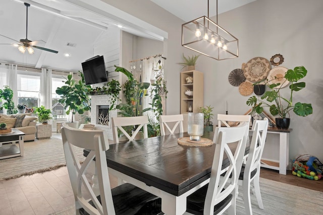 dining room featuring high vaulted ceiling, light hardwood / wood-style flooring, beamed ceiling, ceiling fan, and a fireplace