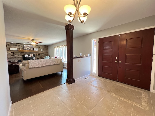 foyer featuring light tile patterned floors, decorative columns, brick wall, a brick fireplace, and ceiling fan with notable chandelier