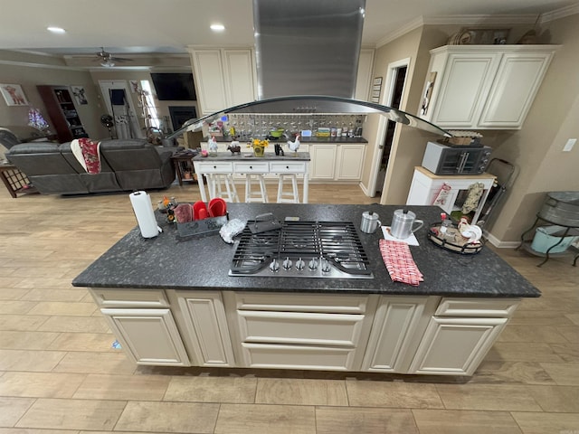 kitchen with stainless steel gas stovetop, dark stone counters, island exhaust hood, ornamental molding, and ceiling fan