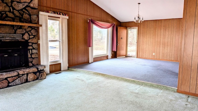 unfurnished living room featuring a notable chandelier, a stone fireplace, vaulted ceiling, wood walls, and light colored carpet