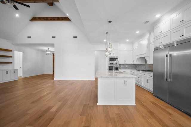 kitchen with built in appliances, light stone countertops, an island with sink, white cabinets, and decorative light fixtures