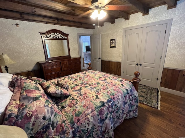 bedroom with dark wood-type flooring, wood walls, a closet, beamed ceiling, and ceiling fan