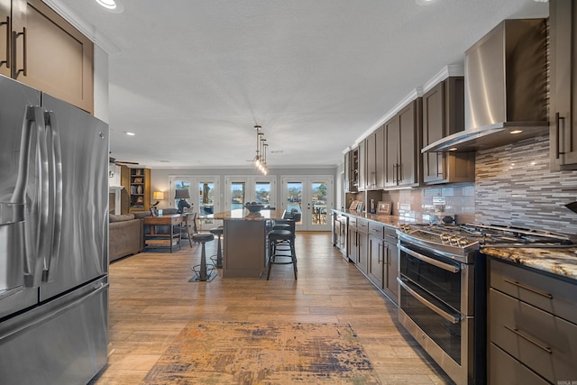 kitchen featuring a breakfast bar area, stainless steel appliances, dark brown cabinets, french doors, and wall chimney range hood