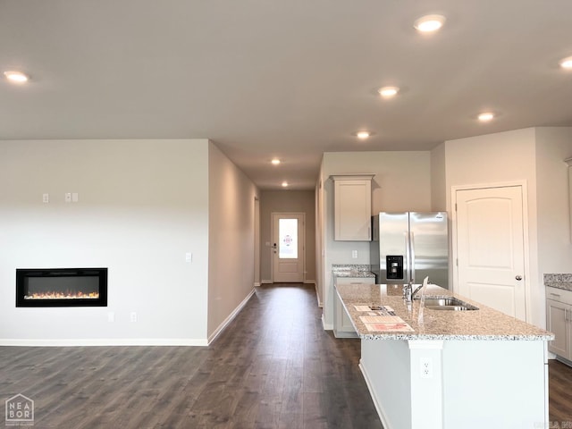 kitchen featuring light stone counters, dark wood-style floors, a center island with sink, a sink, and stainless steel fridge