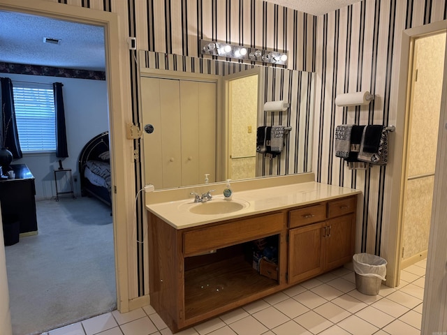 ensuite bathroom featuring wallpapered walls, visible vents, tile patterned floors, a textured ceiling, and vanity