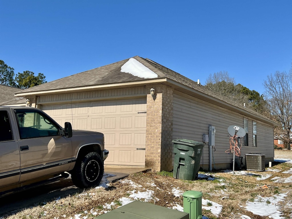 view of snow covered exterior featuring brick siding, an attached garage, cooling unit, and a shingled roof