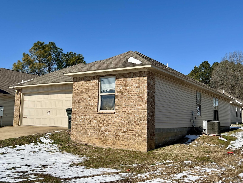 view of snowy exterior featuring an attached garage, driveway, central AC unit, and brick siding