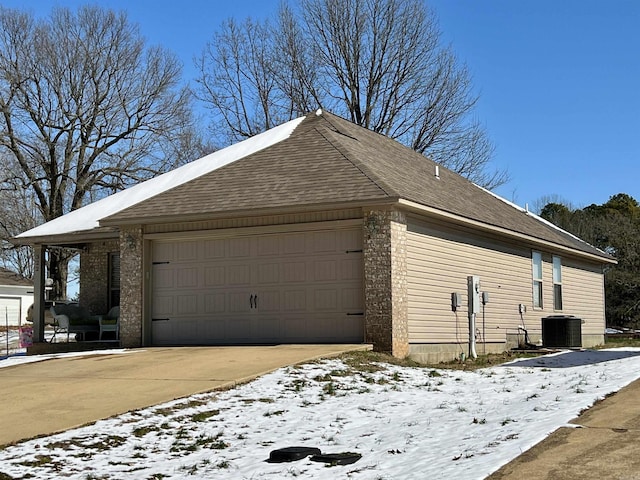 snow covered property featuring a garage, concrete driveway, brick siding, and central AC