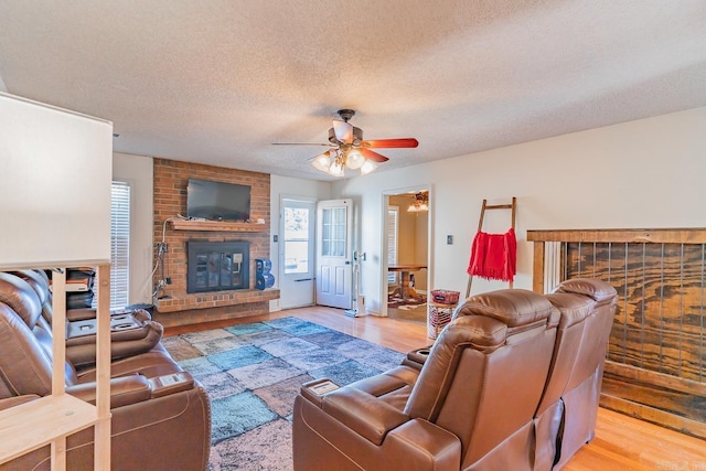 living room featuring light wood-type flooring, a brick fireplace, ceiling fan, and a textured ceiling