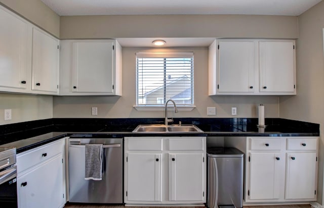 kitchen with stainless steel dishwasher, dark countertops, a sink, and white cabinetry