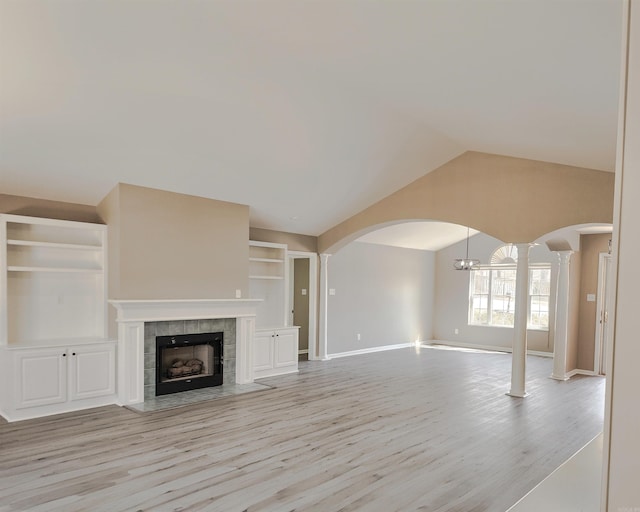 unfurnished living room with arched walkways, built in features, a tile fireplace, vaulted ceiling, and light wood-style floors