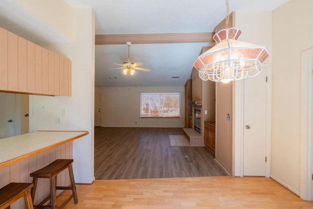 kitchen with light wood finished floors, a glass covered fireplace, ceiling fan, a breakfast bar area, and vaulted ceiling with beams