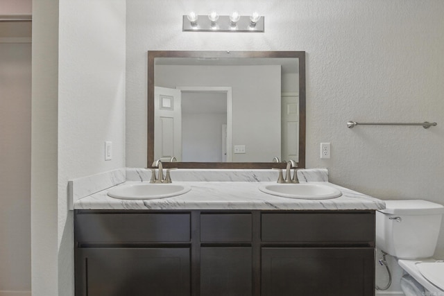 bathroom with a textured wall, double vanity, a sink, and toilet